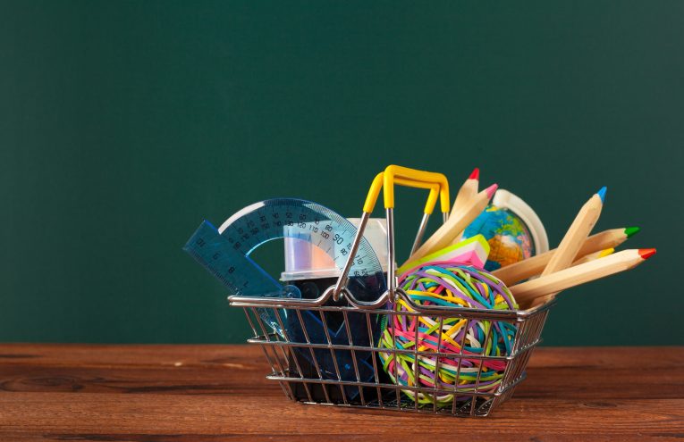 Panier de fournitures scolaires (crayons de couleur, règle, rapporteur, petit globe) devant un tableau vert